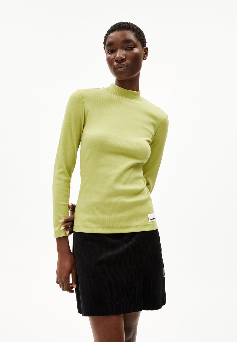 Langsleeve, geribbelde, limoengroene top met een hoge halslijn. Gecombineerd met een zwarte corduroy minirok en een label aan de zoom.