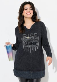 Schwarzes langärmliges Shirt mit Henley-Ausschnitt, das "RISE SHINE" aus Strasssteinchen zeigt. Das Model hält einen mehrfarbigen, strukturierten Trinkbecher.