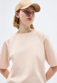 Νεαρό άτομο που φοράει μπεζ καπέλο BOSS και αντίστοιχο ανοιχτό ροζ t-shirt BOSS, κοιτάζοντας ελαφρώς προς τα δεξιά σε ένα απλό φόντο.