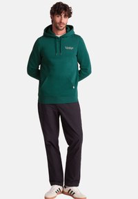 Green hooded sweatshirt in soft fabric with front pouch pocket, adjustable drawstrings, and logo detail on the chest. Paired with dark pants.