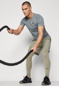 Grå atletisk t-shirt med ett Nike-logga, kombinerad med ljusgröna shorts och åtsittande leggings, som greppar ett svart battle rope, på en enfärgad bakgrund.
