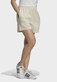 Krämfärgade bomullsshorts med elastisk midja, sidofickor och avslappnad passform, kombinerade med klassiska vita sneakers med svarta ränder.