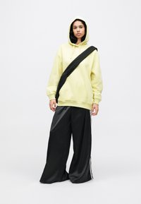 Žltá nadrozmerná mikina s predným vreckom a nastaviteľnou kapucňou, v kombinácii s širokými čiernymi saténovými nohavicami s bočnými pruhmi. Čierna crossbody taška.