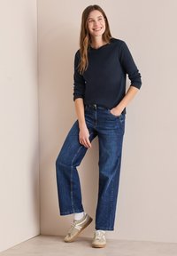 Marinblå långärmad topp, avslappnad passform, tillsammans med blå vida jeans. Tan sneakers med vita accenter. En enkel bakgrund i neutrala färger.