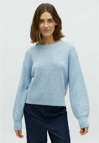 Maglione azzurro chiaro a costine con scollo rotondo e spalle scese. Texture morbida, maniche lunghe e orlo corto. Indossato con pantaloni scuri.