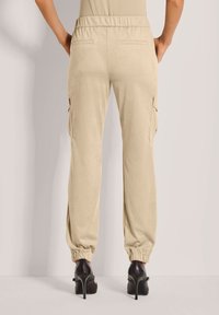 Beige cargo broek gemaakt van zachte stof, met een elastische tailleband, zijzakken en elastische boorden. G gedragen met hoge hakschoenen.