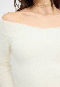 Schulterfreies weißes Sweatshirt aus weichem, flauschigem Material mit subtiler Rippstruktur. Hat einen weiten Ausschnitt und schmal zulaufende lange Ärmel.