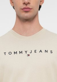 Tricou din bumbac bej, cu inscripția „TOMMYJEANS” brodată în negru, având un mic detaliu logo în roșu, alb și albastru sub text.