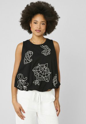 Femme avec coiffure afro portant un haut noir sans manches avec broderie florale blanche et pantalon blanc à cordon sur fond gris.