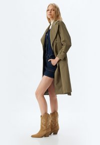 Oliwkowy trench coat o luźnym kroju, noszony na ciemnoniebieski kombinezon z denimu; zestawiony z brązowymi zamszowymi kowbojkami z białymi szwami.