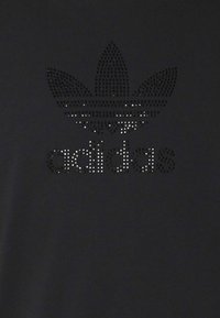 Černé tričko s logem Adidas zdobeným rhinestony a designem trojlístku, umístěným na hrudi, na hladkém povrchu.