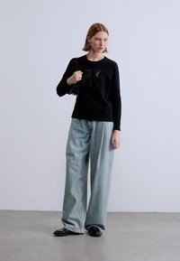 Maglione nero a maniche lunghe con decorazioni lucenti, abbinato a jeans azzurri a gamba larga e scarpe nere. Sfondo uniforme.