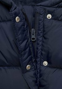 Närbild av en marinblå kviltad jacka med dragkedjan delvis öppen och tre silverfärgade tryckknappar längs framsidan.