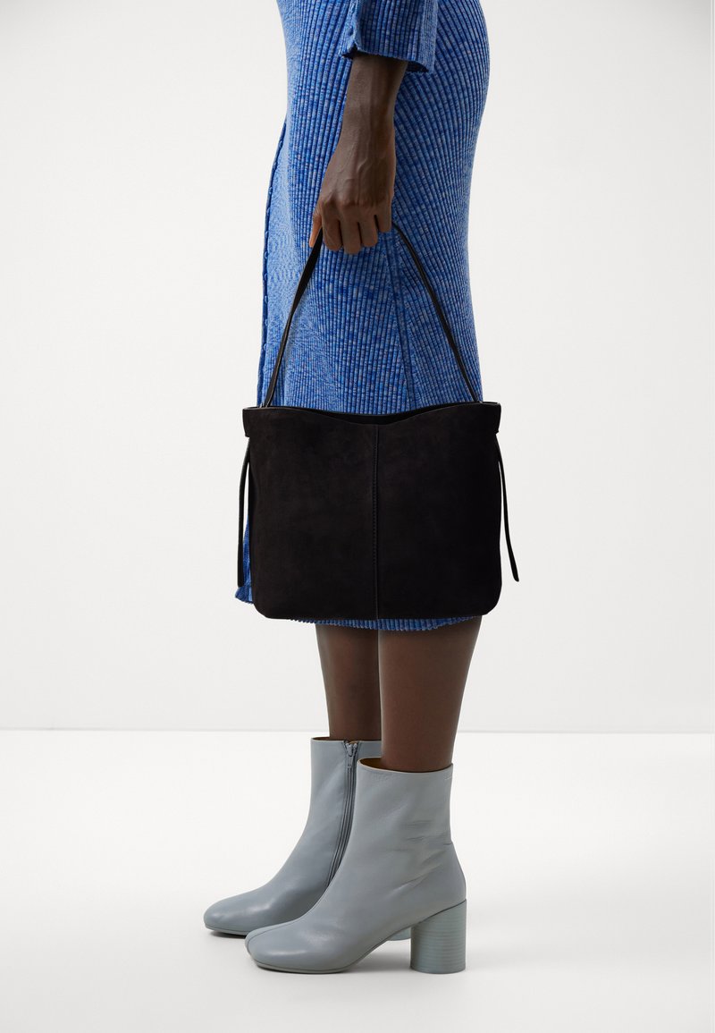 Borsa tote nera in suede con due lacci laterali, tenuta da una persona che indossa un vestito blu texturizzato, abbinata a stivaletti grigi chiari con tacco a blocco.
