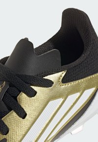 Zapatilla deportiva negra y dorada con un upper texturizado, rayas blancas y un diseño elegante con un collar y cordones acolchados.