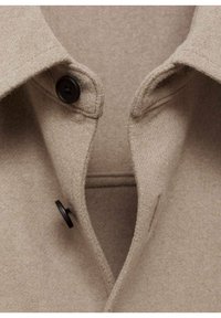 Beige wollen jas met een kraag, voorzien van twee zwarte knopen en een zachte, gestructureerde stof. Zichtbare stiksels bij de kraag.