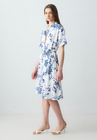 Krótkosleeve, sukienka do kolan z białego materiału w niebieskie kwiatowe wzory, z kołnierzykiem, wiązaniem w talii i zwiewnym dołem.