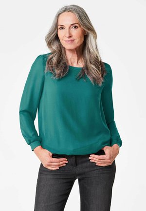 Langärmlige, durchsichtige Bluse in Teal mit Rundhalsausschnitt und gerafften Manschetten; kombiniert mit dunklen Jeans, posiert das Modell mit den Händen in den Hüften.