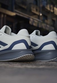 Paar weiße und graue Sportschuhe von hinten auf einer strukturierten Oberfläche mit unscharfem städtischem Hintergrund.