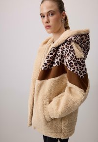 Chaqueta de sherpa color crema con capucha de estampado de leopardo, detalles en marrón y un corte relajado. Cuenta con bolsillos delanteros y un tejido texturizado.