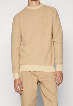Maglione lavorato a maglia beige con motivo testurizzato, collo a giro, polsini e orlo a costine. Indossato sopra pantaloni beige chiaro. Dettaglio logo discreto sul petto.