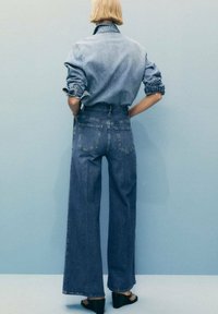 Denim košulja i hlače visokog struka širokih nogavica u srednje plavoj boji. Hlače imaju stražnje džepove i sirovi rub. Model nosi crne platforme.