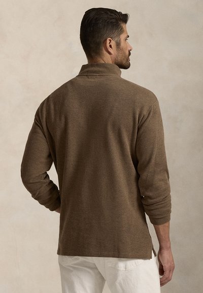 Brun långärmad skjorta med hög krage, texturerad tyg, sidoflikar och figurnära ärmar, bärs över vita byxor.