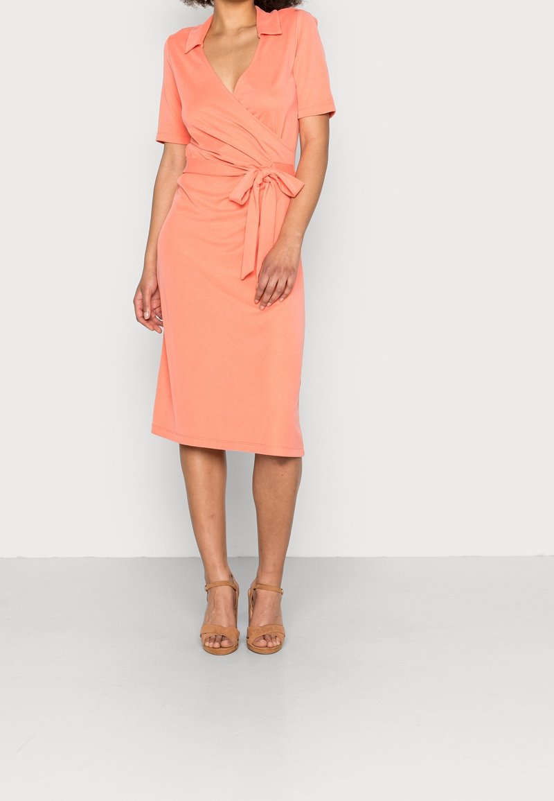 Žena ve korálových šatech s krátkým rukávem a uvázaným pasem, doplněná béžovými sandály na otevřené špičky s kotníkovým páskem, stojící na světlém pozadí.