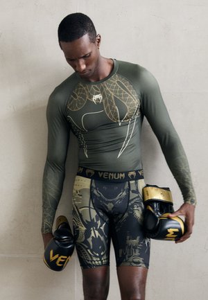 Kompressionsshirt in olivgrün mit Schlangendesign, kombiniert mit gemusterten Shorts. Schwarze Boxhandschuhe mit gelben Akzenten in der Hand gehalten.