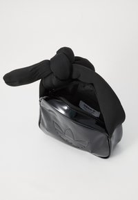 Bolsa preta com material brilhante, apresentando uma forma arredondada. A alça é feita de tecido, com um nó na parte de cima, e possui um logótipo em relevo na parte da frente.