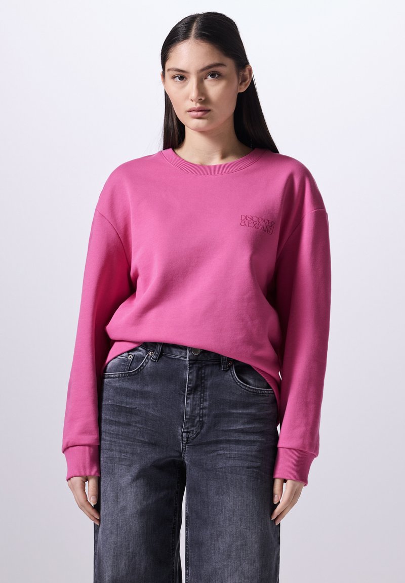 Sudadera rosa con puños y cuello acanalados, que presenta un pequeño logo de "Descubre y Expande". Combinada con unos vaqueros de pierna ancha en gris oscuro.