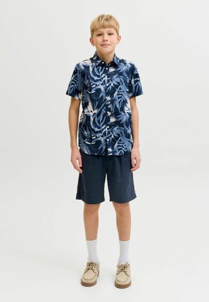 Blond chłopiec stoi, ubraney w niebieską koszulę z krótkim rękawem w wzory, ciemne szorty, białe skarpetki i beżowe buty na jednolitym jasnym tle.