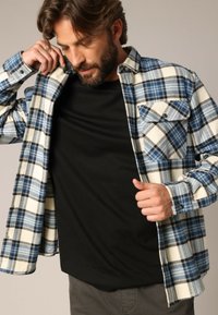 Blauw-wit geruite flanellen shirt met een knoopzak, gedragen open over een zwart T-shirt. Zachte textuur, lange mouwen en kraagontwerp.