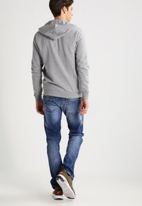 Grå hettegenser med baklomme, sammen med blå jeans. Jeansene har en falmet tekstur, og skoene er mørkegrønne med oransje detaljer.