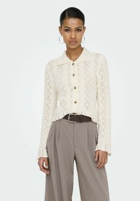 Cardigan crème tricoté avec un col à revers, fermeture boutonnée à l'avant et un motif ajouré complexe, associé à un pantalon beige taille haute et une ceinture marron.