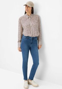 Lichtbeige knoopshirt met donkere stippen, aansluitende blauwe jeans en lichtgekleurde instapschoenen. Accessoires zijn een beige pet.