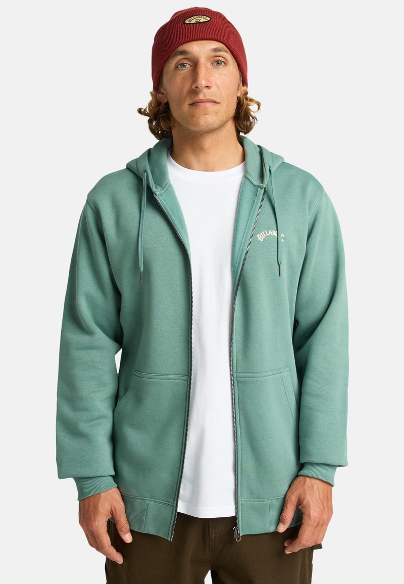 Felpa con zip color teal, con logo bianco sul petto, tasche frontali e cappuccio con cordini. Indossata sopra una T-shirt bianca.