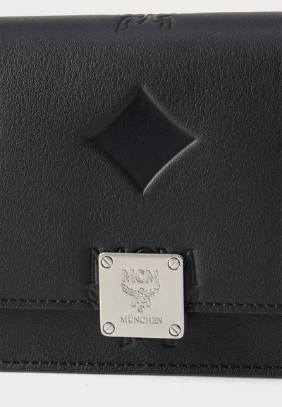 Μαύρο δερμάτινο πορτοφόλι με ανάγλυφο σχέδιο διαμαντιού και ασημί μεταλλική πλάκα με χαραγμένο το λογότυπο "MCM München" στο εμπρόσθιο καπάκι.