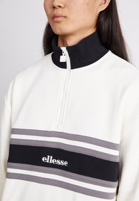 Fleece pullover in crème met een zwarte ribgebreide kraag, voorzien van een halve rits en grijze en witte horizontale strepen, met het logo "ellesse."