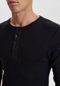 Svart langermet henley-skjorte laget av ribbestrikket stoff, med rund hals og knappestolpe med tre svarte knapper.