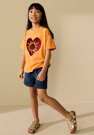 Tüdruk kõnnib toas, seljas oranž T-särk punase südame ja päevalillekujundusega, teksapüksid ja kuldsed sandaalid.