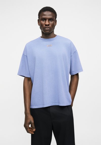 Νέος άνδρας με φαρδιά γαλάζια μπλούζα και μαύρο παντελόνι, που στέκεται μπροστά σε απλό λευκό φόντο.