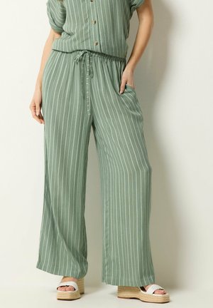 Kvinde iført løse, grønne bukser med striber og snøre i taljen, samt hvide platformssandaler, stående med den ene hånd i lommen.