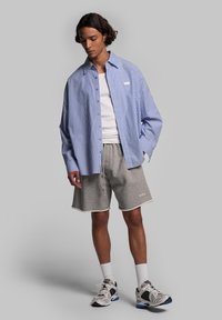 Camicia oversize a righe blu e bianche, pantaloni corti grigi, calzini bianchi e scarpe da ginnastica nere e bianche. Stile casual, vestibilità rilassata.