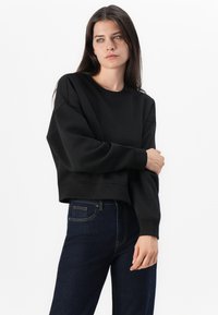 Felpa nera corta con maniche lunghe, scollo rotondo e polsini a coste, abbinata a jeans blu scuro. Design semplice e pulito.