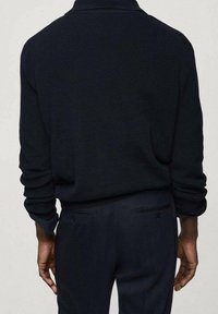 Maglione a coste di un blu navy scuro con colletto aderente e maniche lunghe, abbinato a pantaloni scuri su misura con tasche a filetto.
