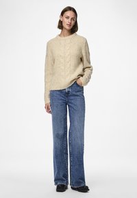 Camisola bege de malha com padrão de trança e detalhes canelados, combinada com jeans azuis flare. O tecido tem uma aparência texturada e aconchegante.