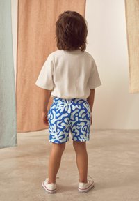 Enfant portant un T-shirt blanc ample et un short bleu à motifs avec des formes abstraites blanches, chaussé de baskets blanches, de dos contre des fonds en tissu.