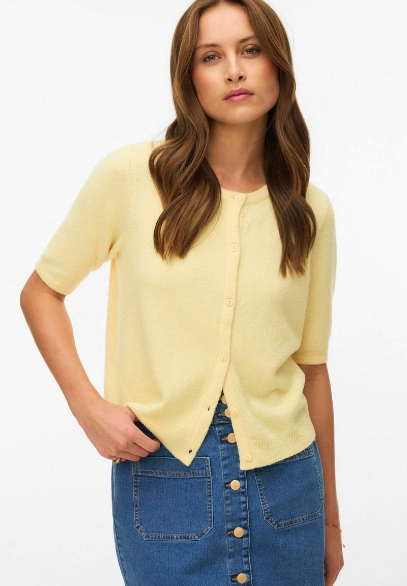 Kvinde med langt brunt hår iført en bleg gul kortærmet knap-cardigan og et blåt denimskørt med knapper foran, poserer med den ene hånd i lommen.
