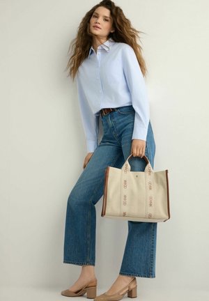 Kobieta ubrana w jasnoniebieską koszulę na guziki, niebieskie wysokie spodnie dżinsowe, beżowe buty na blokowym obcasie, trzyma beżową torbę płócienną z brązową lamówką.